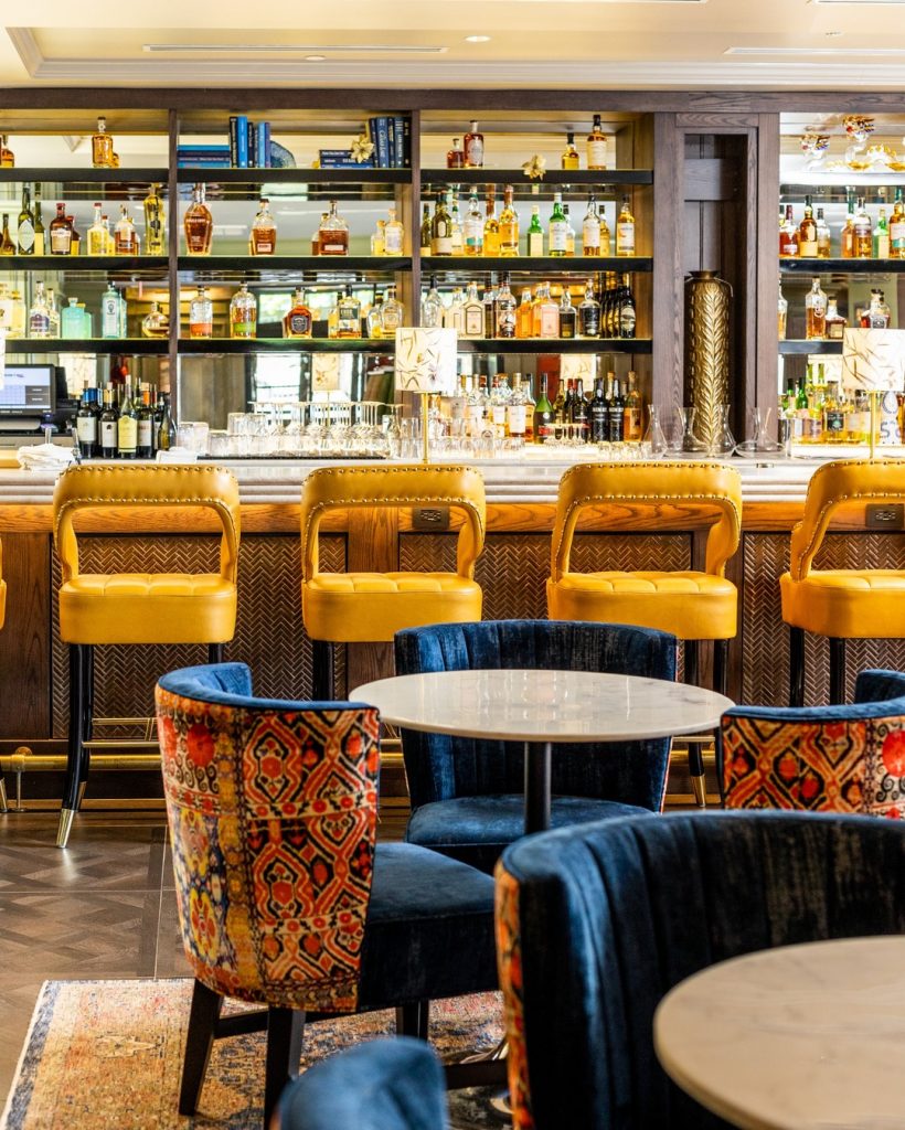 Interior shot of restaurant furnished with yellow and blue, upholstered chairs and a well stocked bar at FIA Restaurant & Bar