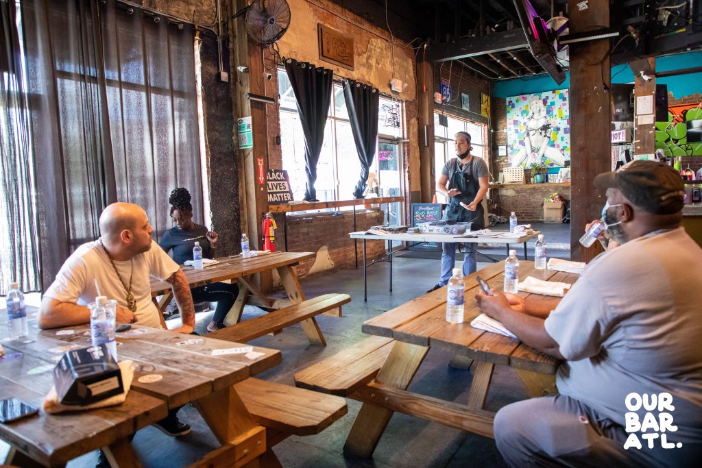 Guests at an open plan bar with curtain covered windows, wooden benches and tables and fans hanging from brick walls at Our Bar ATL