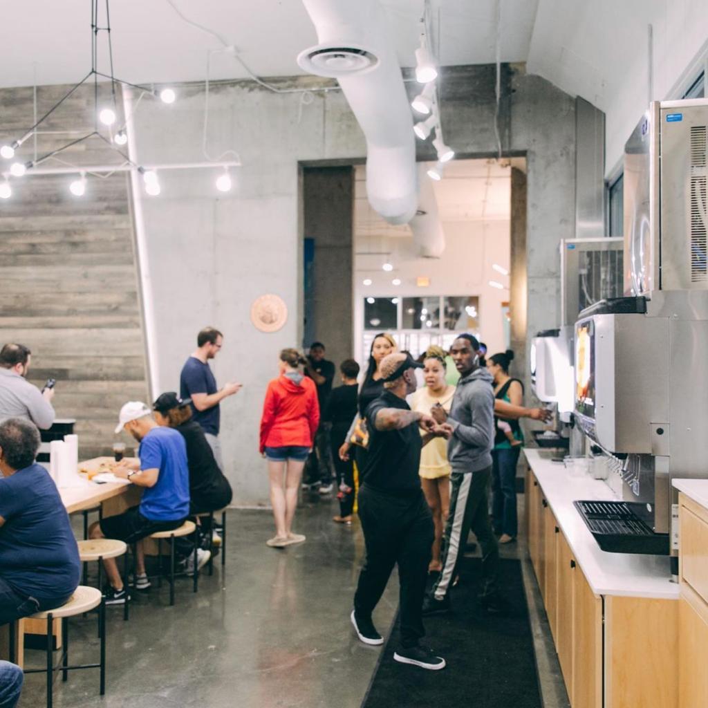 Patrons milling about a pizzeria with pale gray walls, white lights and brown stools at Slim & Husky’s Pizza Beeria